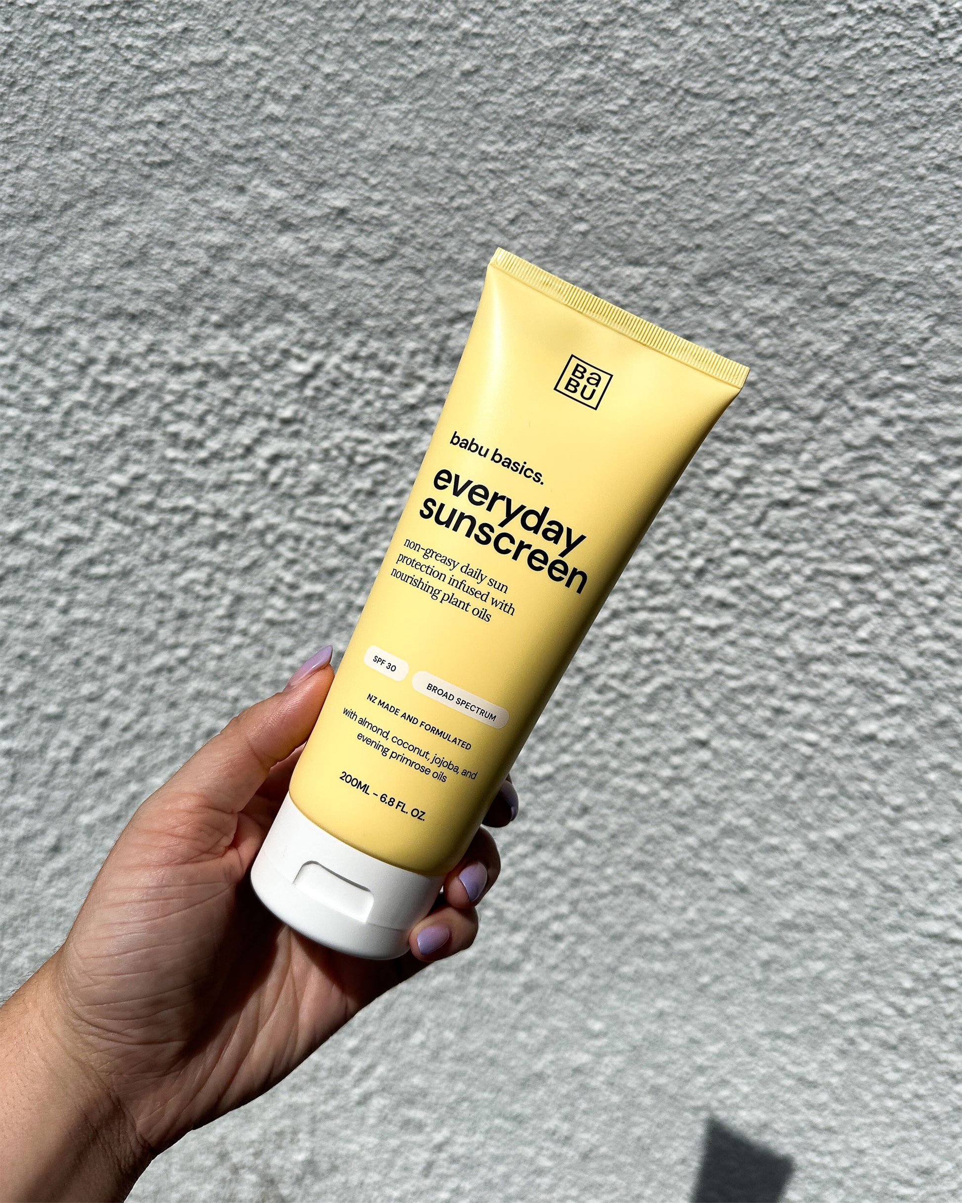 Hand holding a tube of Babu Basics everyday sunscreen against a grey background