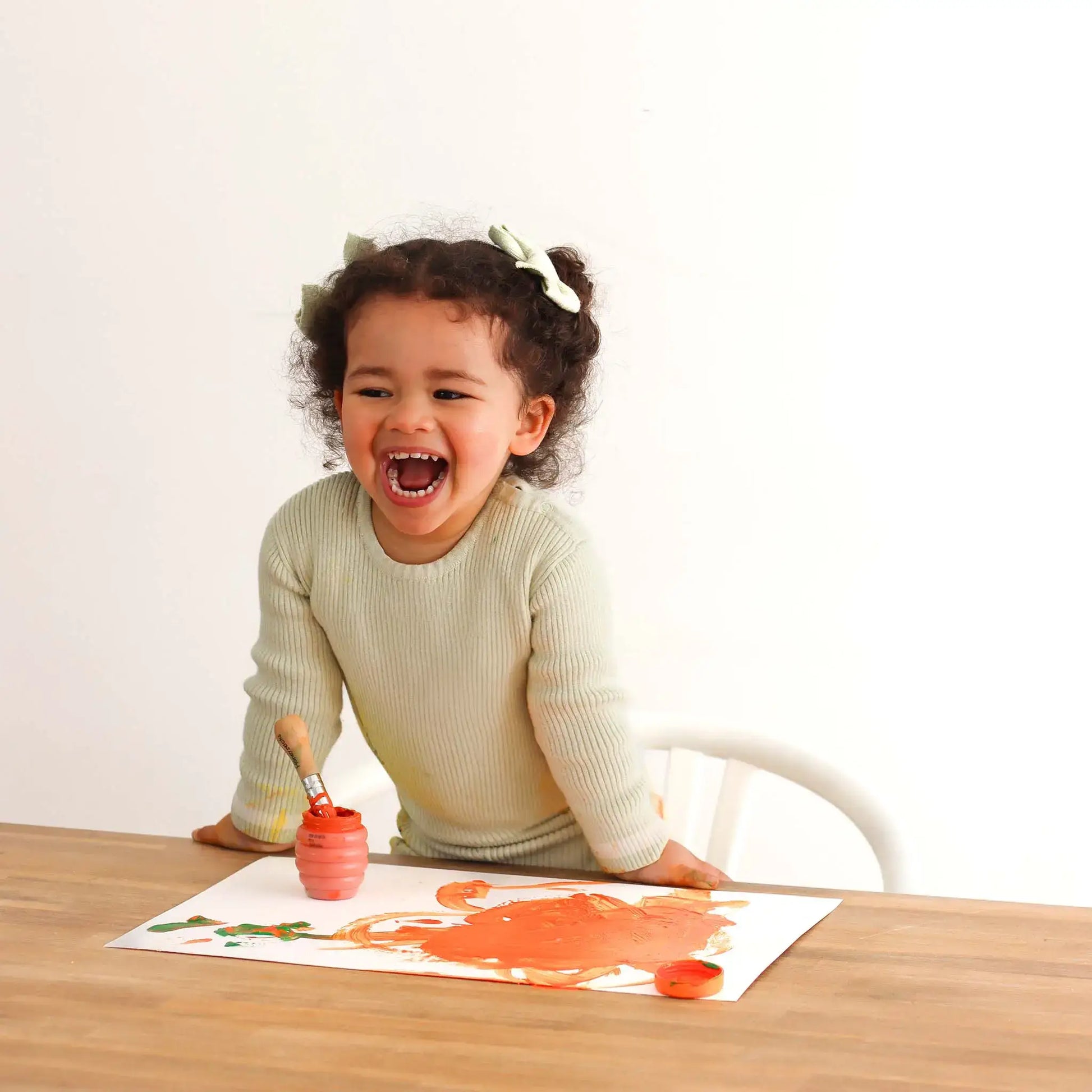 Child painting on a paper with a brush and container of paint, sitting at a table.