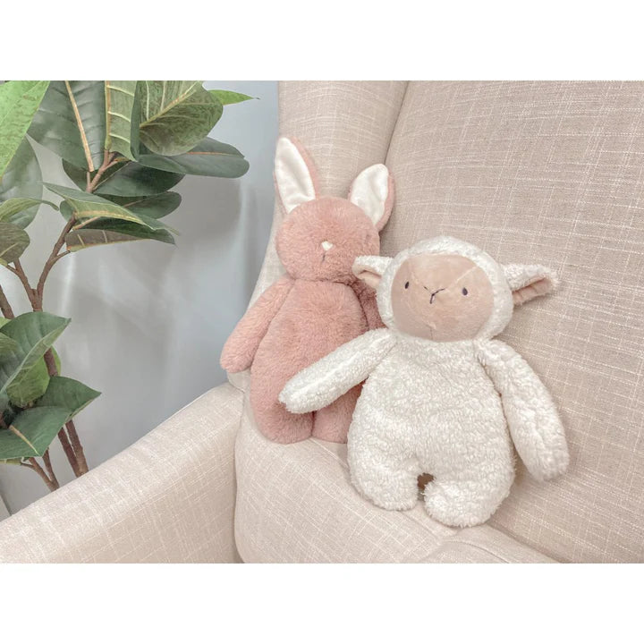 Two plush toys, one pink and one white, sitting on a light-colored surface with a plant in the background.