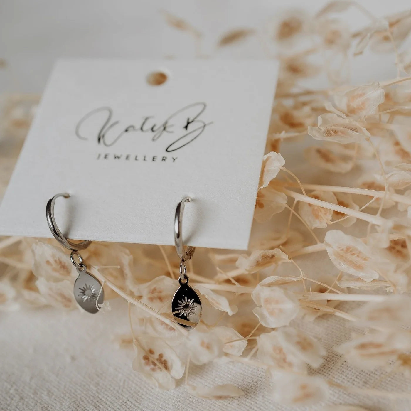 Silver earrings on a white card with 'KatyB Jewellery' branding, placed on dried flowers.
